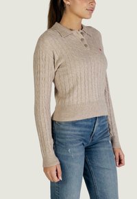 Beige kabelgebreide trui met een kraag en drie knopen, voorzien van geribbelde manchetten en zoom. Gecombineerd met een blauwe spijkerbroek.