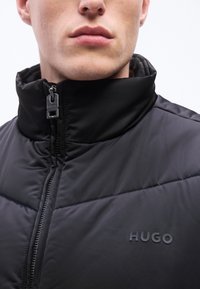 Κοντινό πλάνο ενός ατόμου που φοράει μαύρο μπουφάν τύπου κοντό μπουφάν-πουφ με ανεβασμένο γιακά και λογότυπο "HUGO" στο στήθος, με το φερμουάρ μερικώς ανοιχτό.