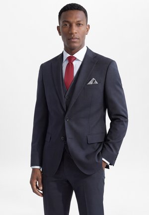 Man die een donker marineblauw pak draagt met bijpassend gilet, wit overhemd, rode stropdas en grijs pochet, staat met één hand in de zak.