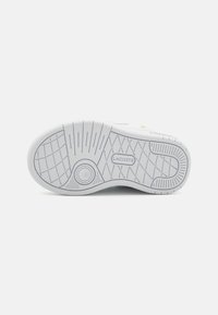 Semelle de chaussure de sport blanche avec un motif de grip texturé, comportant un accent circulaire et une étiquette de marque au centre. Matériau en caoutchouc lisse.