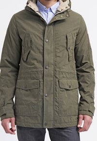 Olive green parka jacket with a beige hood, two front pockets, snap buttons, and a cinched waist. Textured fabric with a casual design.