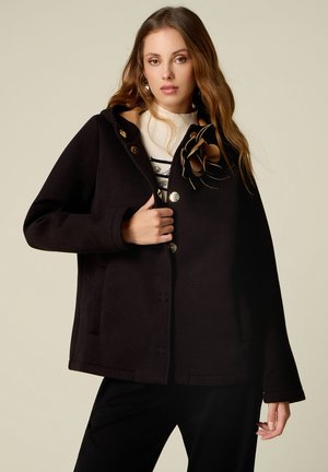 Young woman with long hair wearing a black buttoned coat with a large flower detail and a light turtleneck underneath.