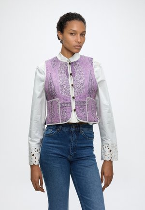 Frau trägt eine weiße Bluse mit Spitzenmanschetten, eine lila gemusterte, geknöpfte Weste und blaue hoch taillierte Jeans vor einfarbigem Hintergrund.