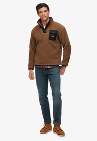 Brown fleece pullover with a high collar and black chest pocket, paired with blue jeans and brown boots. Textured, warm material.