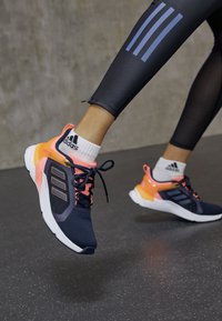 Des chaussures de sport avec une tige en maille bleu marine, des accents roses et oranges, une semelle blanche et des lacets noirs ; portées avec des leggings noirs texturés.