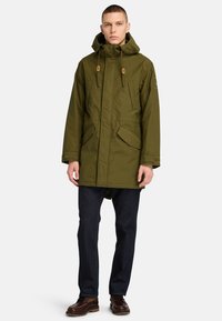 Olivgrön parkas med huva, framfickor, justerbara snörningar och dragkedja, kombinerad med mörk denim och bruna stövlar.