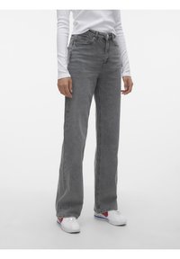 Mujer con vaqueros de tiro alto y pierna recta en gris, camiseta blanca de manga larga y zapatillas blancas con detalles en rojo y azul, parada sin moverse.