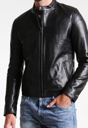 Hombre con una chaqueta de cuero negra ajustada con bolsillos con cremallera y vaqueros azules, mano casualmente en el bolsillo, sobre un fondo liso.