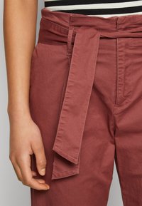 Rotbraune Hose mit breitem Bund und gebundenem Gürtel. Aus glattem Stoff gefertigt, mit Seitentaschen und lässigem Schnitt.