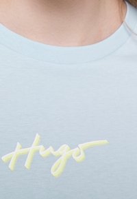 Světle modré bavlněné tričko s bílým a žlutým kurzívním logem "Hugos". Tričko má kulatý výstřih s žebrovaným lemem.