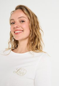 Camiseta blanca de algodón con un diseño de "Amor" bordado en oro. Cuello redondo, mangas cortas y acentos de botones en los laterales. Textura suave.