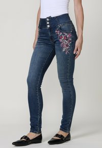 Vaqueros skinnies de color azul oscuro con cintura alta, que presentan un bordado floral decorativo en la cadera izquierda y un cierre de botón metálico en la parte delantera.