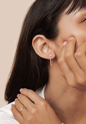 Gold earrings with delicate chain and bead accents, worn on an ear, with a close-up of smooth skin and light brown hair. Pink manicure visible.