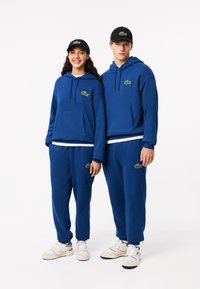Blauwe katoenen hoodies en sweats met kangoeroezakken, met geborduurde groene krokodillogo's. Zwarte petten maken de outfits compleet.