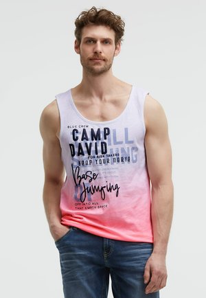 Mann mit lockigem Haar, trägt ein ärmelloses weiß-rosa Ombre-Top mit Schriftzug und blaue Jeans, steht mit einer Hand in der Tasche.