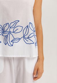 Top de lino blanco con diseño de hojas bordado en azul, que cuenta con un corte holgado y una textura suave. El dobladillo es recto y tiene un acabado minimalista.
