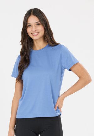 Femme souriante aux longs cheveux foncés portant une chemise bleu clair à manches courtes et un pantalon noir, debout, une main sur la hanche, sur fond blanc.