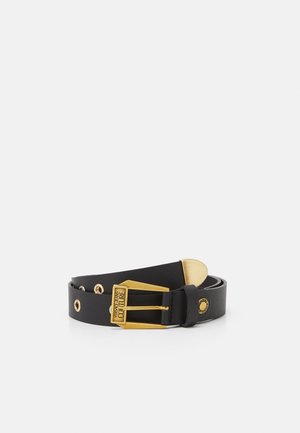 Czarny skórzany pasek ze złotą klamrą z wygrawerowanym napisem „Versace Jeans Couture” i złotymi przelotkami, zwinięty na białej powierzchni.
