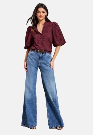 Donna che indossa una blusa color bordeaux con maniche a sbuffo a occhielli, infilata in jeans a gamba larga a vita alta, con cintura marrone e sandali neri.