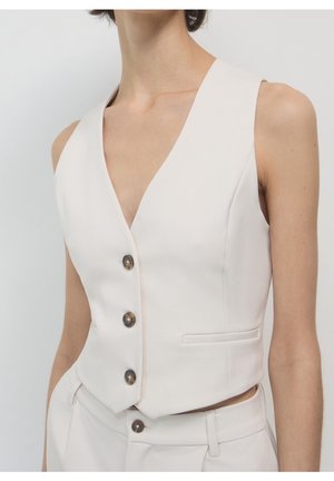 Gilet blanc sans manches, taillé sur mesure avec un décolleté en V profond et quatre boutons, porté avec un pantalon taille haute assorti sur un fond uni.