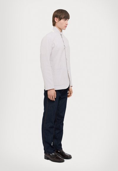 Hellgraue, strukturierte Jacke mit Stehkragen, Knopfleiste und seitlichen Taschen, kombiniert mit dunklen Jeans und schwarzen Schuhen.