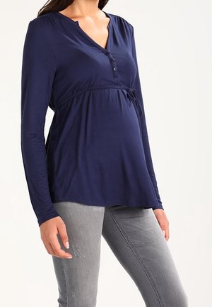 Femme enceinte portant un haut de maternité bleu marine à manches longues avec des boutons et un jean gris ajusté, debout devant un fond clair uni.