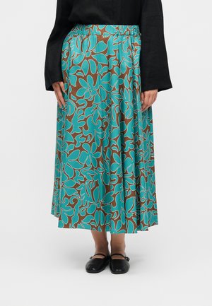 Person iført en midilængde nederdel med turkis og brunt blomsterprint, sort langærmet top og sorte Mary Jane-flade sko, stående mod en hvid baggrund.