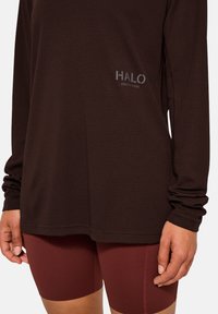 Braunes langärmliges Feuchtigkeitsregulierendes Shirt aus strukturiertem Stoff mit einem Logo. Kombiniert mit bordeauxroten figurbetonten Shorts, die Nahtdetails zeigen.