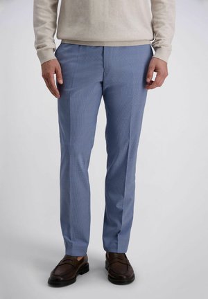 Man draagt lichtblauwe op maat gemaakte broek, beige trui en donkerbruine leren loafers, staand tegen een effen achtergrond.