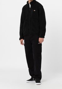 Zwarte fleecjas met ritszakken, hoge kraag en textuurstof. Gecombineerd met zwarte cargobroek en zwarte sneakers.