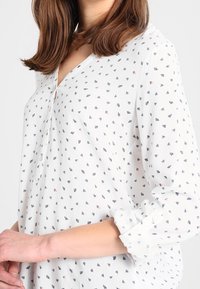 Blusa bianca con scollo a V e piccoli motivi triangolari multicolori. Maniche lunghe con polsini a bottone, realizzata in tessuto leggero.