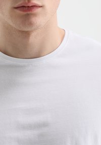 Biely bavlnený tričko s okrúhlym výstrihom, s hladkou textúrou a bez viditeľných vzorov alebo grafiky. Detailný pohľad na výstrih.