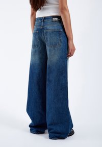Wide-leg blue denim jeans with a faded finish, featuring two back pockets and a branded tag on the waistband.