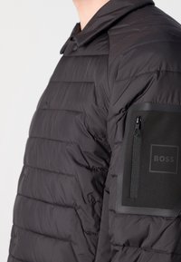Black puffer jacket, quilted design with horizontal lines, features a snap collar and a zippered pocket on the left arm with logo accents.