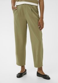 Pantalon vert olive ajusté avec plis sur le devant, poches latérales et coupe décontractée. Porté avec des chaussures plates noires, mettant en valeur la texture lisse du tissu.