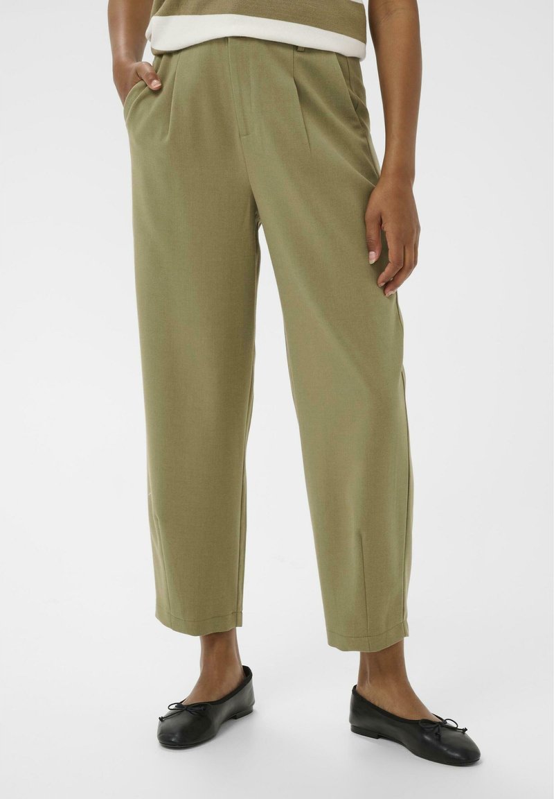 Pantalon vert olive ajusté avec plis sur le devant, poches latérales et coupe décontractée. Porté avec des chaussures plates noires, mettant en valeur la texture lisse du tissu.