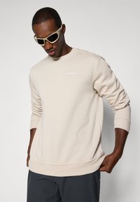 Sweatshirt de algodão bege com gola redonda, mangas longas, punhos canelados e patch de logótipo; usado com óculos de sol de cor clara contra um fundo simples.