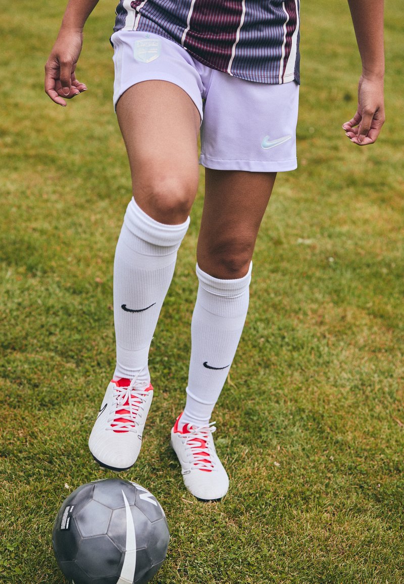 Shorts de football blancs Nike avec une teinte lavande, chaussettes blanches arrivant aux genoux, chaussures blanches avec des accents rouges et un ballon de football noir sur l'herbe.