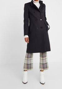 Abrigo negro de doble breasted con botones, dos bolsillos laterales y un corte entallado, usado sobre unos pantalones de cuadros y botas blancas hasta el tobillo.
