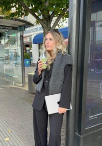 Nainen mustassa puvussa pitää kädessään vihreää juomaa ja Apple-kannettavaa, päällään harmaa pörröinen huivi, seisten bussipysäkin lähellä.