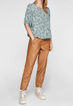 Femme portant un chemisier bleu clair à motifs avec manches mi-longues, pantalon en cuir synthétique beige et baskets plateformes blanches, debout les mains dans les poches.