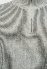 Lysegrå ribbestrikket genser med skjult glidelås midt foran ved halsen og høy krage.