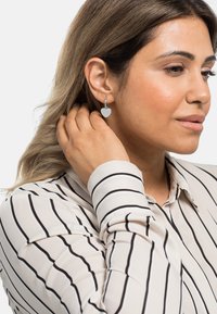 Orecchini a forma di cuore in argento con strisce verticali, attaccati a un cerchio. La modella indossa una blusa a righe con bottoni, mentre si tocca i capelli.