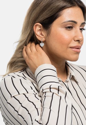 Silberne herzförmige Ohrringe mit vertikalen Streifen, die an einem Ohrstecker befestigt sind. Das Modell trägt eine gestreifte Bluse mit Knöpfen und berührt ihr Haar.