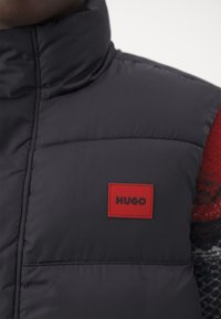 Zwarte puffer vest met hoge kraag, voorzien van een rood HUGO logo patch. Gladde, gewatteerde textuur met horizontale stikseldetails.