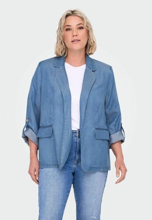 Denim blejzr v světle modré barvě s přiléhavým střihem, s klopami, dvěma předními kapsami a ohrnutými rukávy. Nosený přes bílé tričko a džíny.
