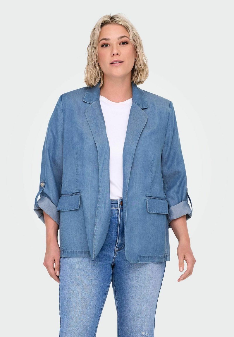 Blazer de mezclilla en azul claro con un corte entallado, solapas con muesca, dos bolsillos delanteros y mangas enrolladas. Usado sobre una camiseta blanca y jeans.