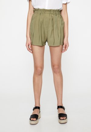 Mujer con pantalones cortos plisados de tiro alto en verde oliva con bolsillos laterales y sandalias negras de plataforma con tiras en el tobillo.