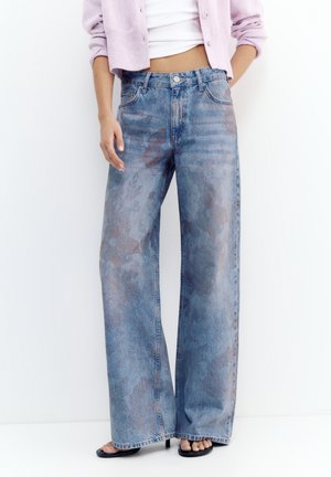 Kvinde iført blå jeans med høj talje og vidde i benene med et falmet mønster, stylet med en kort hvid top og en lyserød cardigan.