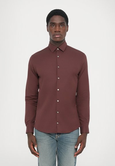 Camisa de manga larga burdeos con cuello abotonado, botones plateados y un corte entallado, combinada con unos vaqueros azul claros. Textura suave.